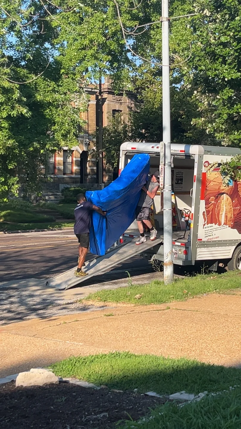 Two crew members moving a fully covered large item up a box-truck ramp